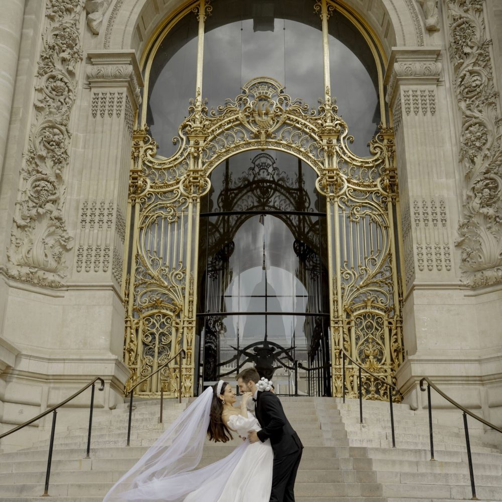Bruid en bruidegom delen een kus op de trappen van een paleisachtig gebouw met gouden poort, haar sluier wappert in de wind. door Lavender Photography. bruiloft fotograaf. trouwfotograaf. fotograaf bruiloft. fotograaf buiten bruiloft. huwelijksfotograaf. wedding fotograaf. loveshoot fotograaf Professionele bruiloftsfotograaf
